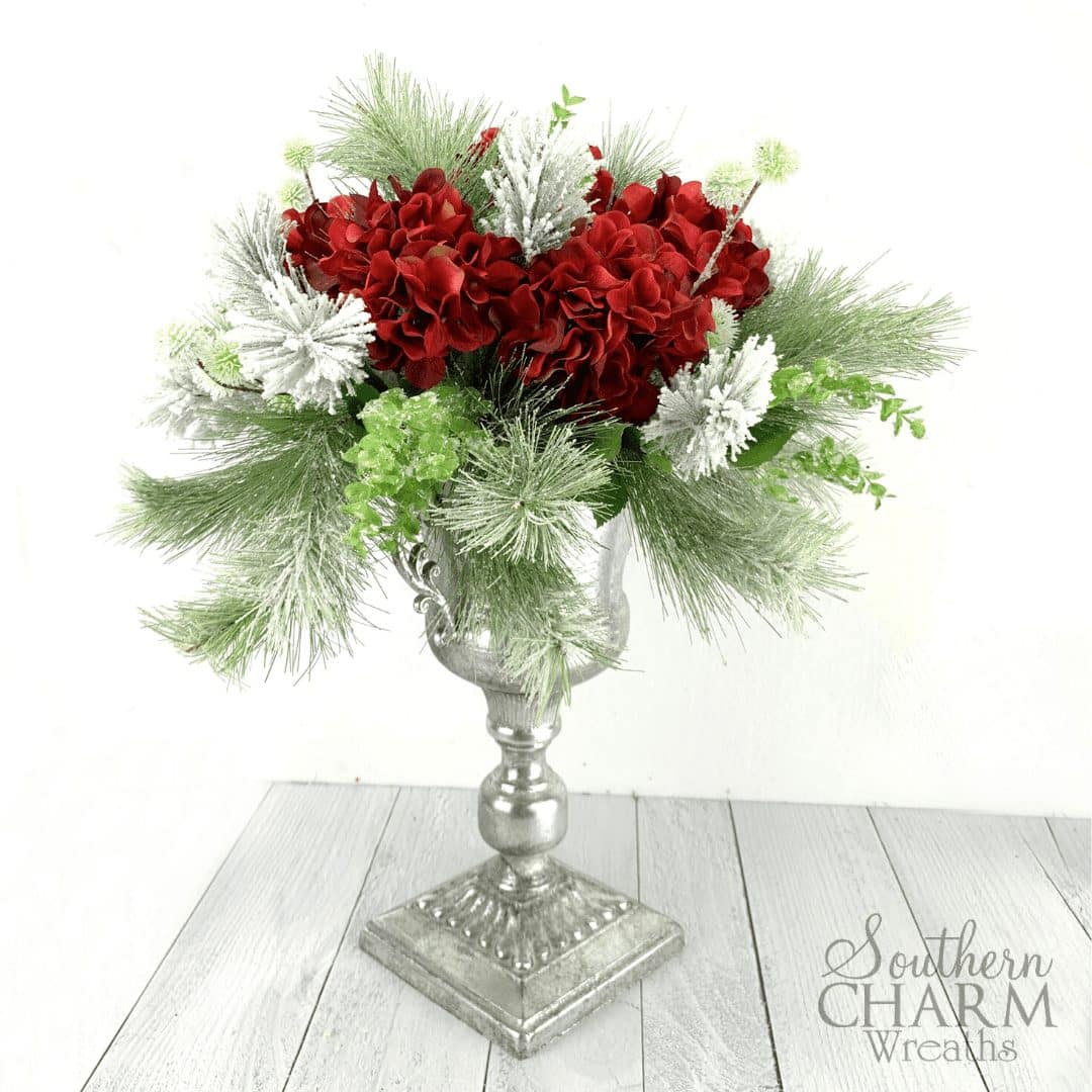 DIY Hydrangea Christmas Centerpiece, Table arrangement with evergreen branches, greenery, and red silk flowers, in a silver pedestal container. In this video, learn to make an elegant Hydrangea Christmas Centerpiece in 5 simple steps.