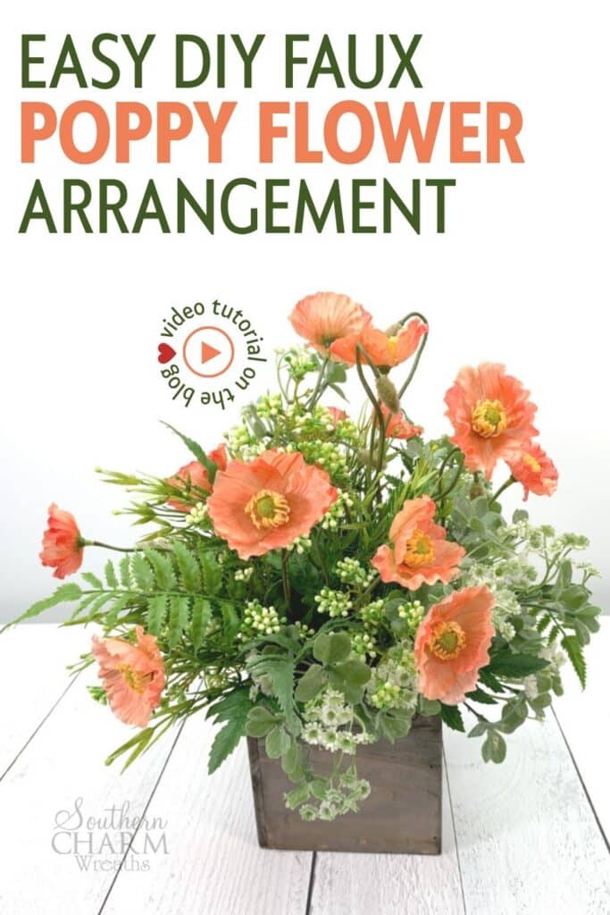 Coral faux poppy floral arrangement in a small wood box container, made with silk poppies, fern, clover, and white buds, sitting on a white wood surface.