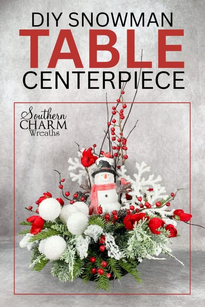 Handmade Christmas table centerpiece featuring a snowman figurine, glittery white snowballs, red berries, frosted greenery, and white snowflake accents. A festive DIY holiday decoration.