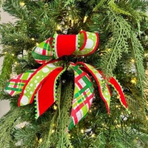 A festive Christmas tree bow made with bright red, green, and plaid wired ribbon, accented with pom-pom trim, perfectly nestled among lush greenery on a decorated tree.