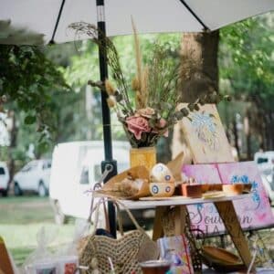 Handmade vendor booth at an outdoor craft show featuring a vase of dried and silk flowers, woven bags, painted artwork, and handmade goods displayed under a white umbrella.