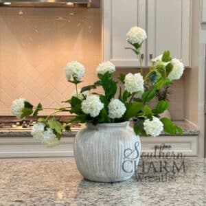 White snowball hydrangeas arranged asymmetrically in a textured ceramic vase on a granite kitchen island with Southern Charm Wreaths watermark.