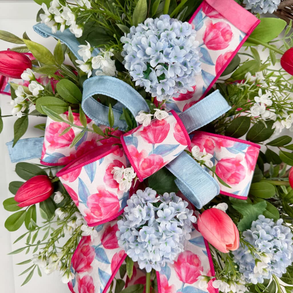 Close-up of a layered spring wreath bow made with pink tulip floral ribbon and soft blue wired ribbon, surrounded by pink tulips, blue hydrangeas, and lush greenery.