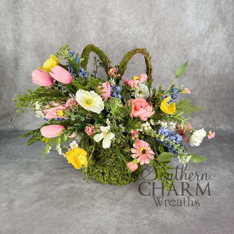 Easter bunny basket silk flower table arrangement with pink tulips, peach ranunculus, white anemones, pink gerbera daisies, blue delphinium, and lush mixed spring greenery cascading from a moss-covered grapevine bunny basket with bunny ear handles