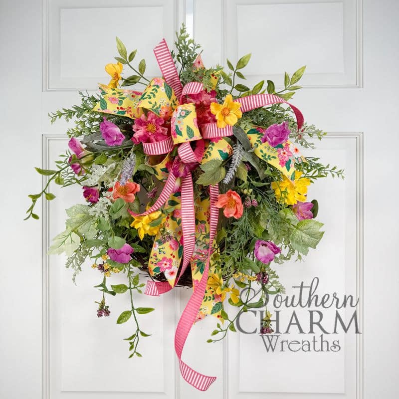 Summer wildflower wreath on a white paneled door featuring layered silk florals in hot pink, yellow, coral, orange, and purple, with trailing mixed greenery and a double-ribbon bow in yellow floral print and pink stripe at the top.