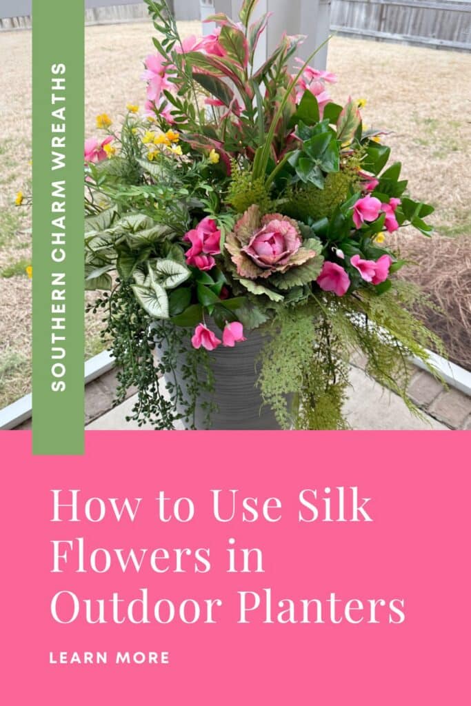 Angled view of a silk flower outdoor planter in a gray ribbed pot. The arrangement features tall pink tropical stems at the back, a silk ornamental kale focal flower at the front center, clusters of pink geraniums and filler blooms, yellow accent flowers, caladium leaves, and trailing string of pearls and wispy greenery over the pot edges. A green vertical banner on the left reads Southern Charm Wreaths in white. A pink footer block reads: How to Use Silk Flowers in Outdoor Planters, with Learn More below.
