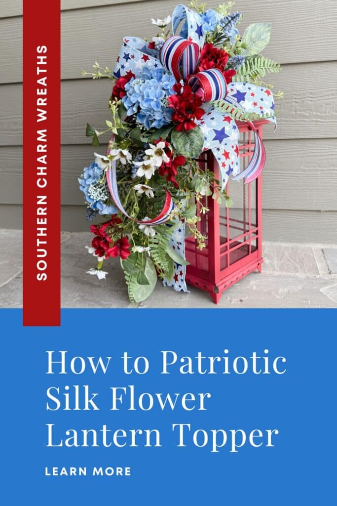 Patriotic silk flower lantern topper with blue hydrangeas, red geraniums, white daisy filler flowers, lavender blooms, fern and salal greenery, and a large star-print patriotic ribbon bow with red-white-blue striped accents, displayed on a red metal lantern on a stone patio — Southern Charm Wreaths how-to tutorial