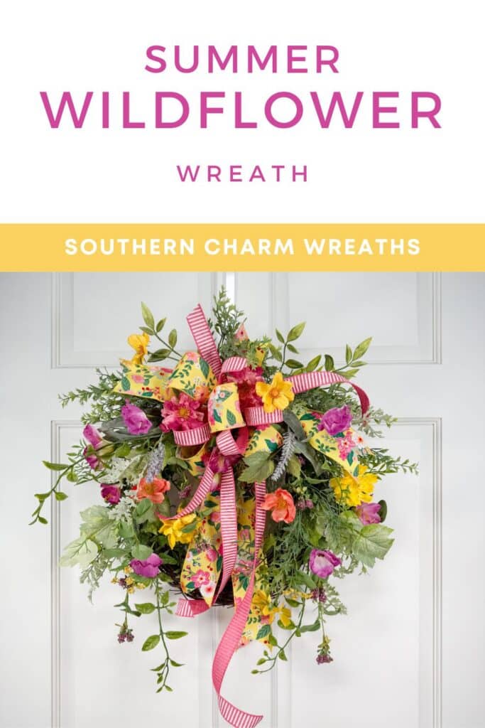Full view of a summer wildflower wreath hanging on a white paneled door. The grapevine base is covered in hot pink, yellow, coral, orange, and purple silk flowers with multiple types of trailing greenery extending well beyond the base edges. A two-ribbon bow in yellow floral print and pink-and-white stripe sits at the top of the wreath. White text header above reads: Summer Wildflower Wreath. Yellow banner below reads: Southern Charm Wreaths in white.