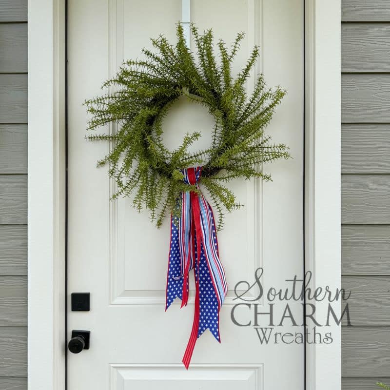 A full green fern wreath hanging on a white paneled front door with gray siding. A patriotic ribbon wreath sash made from three layered wired ribbons — navy blue with white stars, red and white stripe, and light blue and white stripe — is attached at the bottom center of the wreath and drapes in flowing tails below the base.