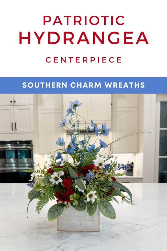 Patriotic table arrangement in a whitewashed wood box on a marble kitchen island. The silk flower arrangement features tall blue cosmos and grass stems at the top, red hydrangeas in the center, white daisy filler, blue muscari sprays, and mixed greenery including variegated nettle leaf and fern. White text header above reads: Patriotic Hydrangea Centerpiece in red. Blue brand bar reads: Southern Charm Wreaths in white.