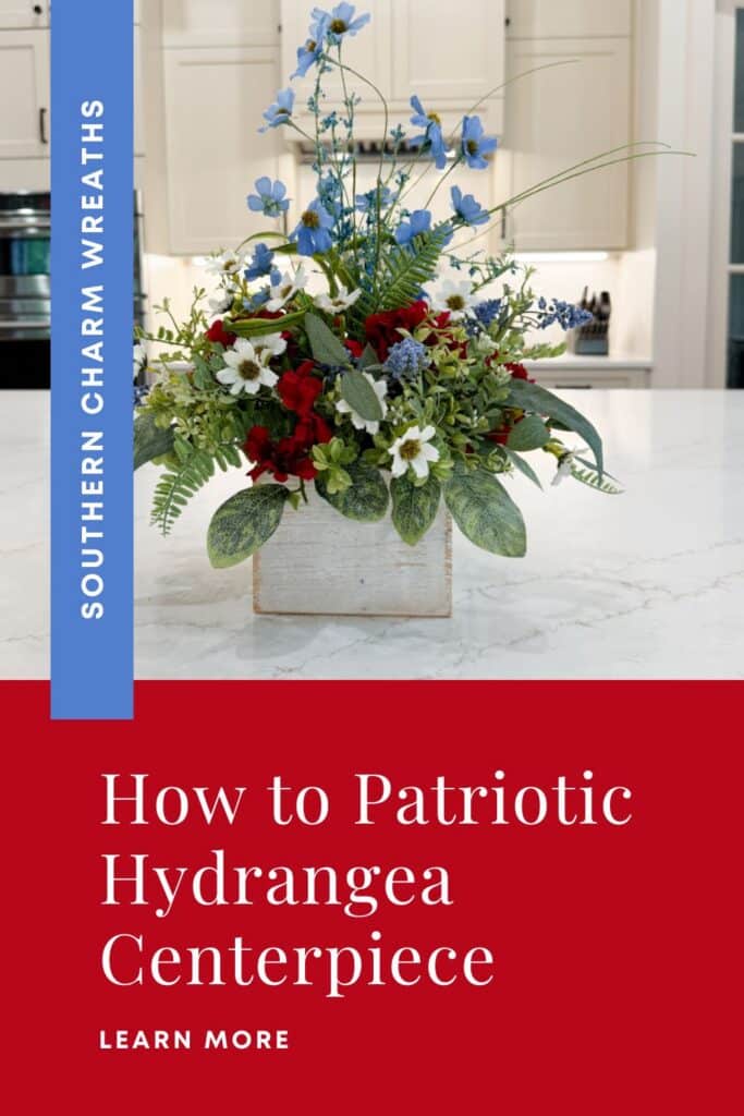 Patriotic table arrangement in a whitewashed wood box on a marble kitchen island. The silk flower arrangement features tall blue cosmos and grass stems at the top, red hydrangeas in the center, white daisy filler, blue muscari sprays, and mixed greenery including variegated nettle leaf and fern. White text header above reads: Patriotic Hydrangea Centerpiece in red. Blue brand bar reads: Southern Charm Wreaths in white.