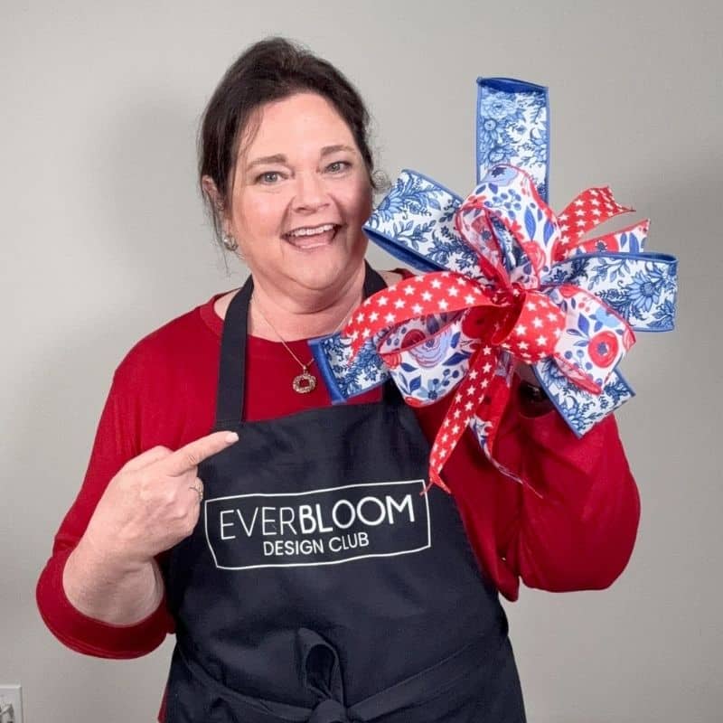 Julie Siomacco of Southern Charm Wreaths smiles and points to her black Everbloom Design Club apron while holding a large patriotic multi-ribbon bow made from blue floral, red star, and white and blue wired ribbons held up beside her shoulder. Neutral gray background. Square format with no text overlay.