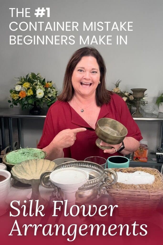 Julie Siomacco of Southern Charm Wreaths smiles and points to a bronze pedestal bowl container she is holding up, with several other containers of varying sizes displayed on the table in front of her including a shell-shaped container with green foam, a silver handled urn, a white pedestal, and a teal mug. Completed silk flower arrangements are visible on the shelving behind her. White text at the top reads: The #1 Container Mistake Beginners Make In. A red footer overlay reads: Silk Flower Arrangements in white script.