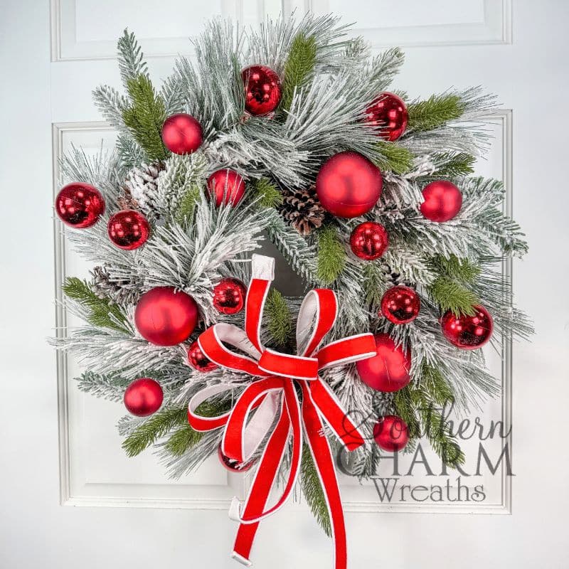 Finished flocked Christmas wreath hanging on a white paneled door, with snowy pine, green pine sprigs, pine cones, shiny and matte red ornaments, and a red velvet bow with white edges. Southern Charm Wreaths logo in the lower right corner.