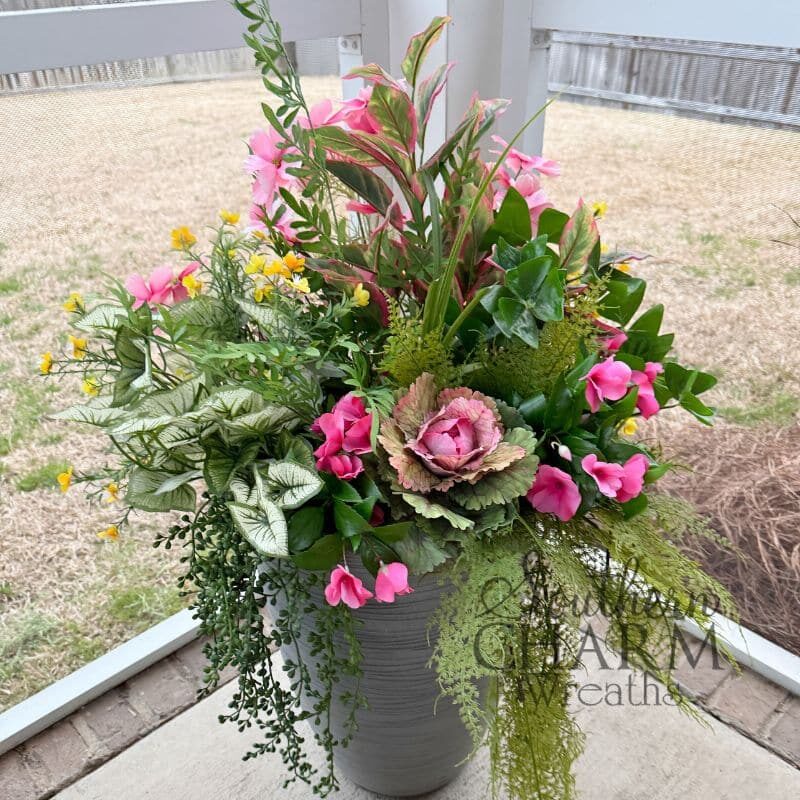Outdoor planter arrangement featuring a silk ornamental kale focal flower, pink geraniums, yellow accent blooms, caladium foliage, and trailing string of pearls greenery in a gray ribbed pot on a covered porch. Southern Charm Wreaths.