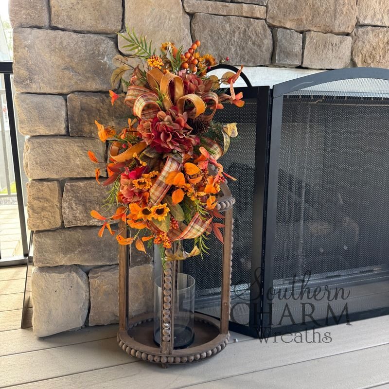 DIY fall lantern swag with rust and burgundy silk hydrangeas, mini sunflowers, orange fall leaves, berries, and a plaid and orange velvet ribbon bow, shown against a stone fireplace.