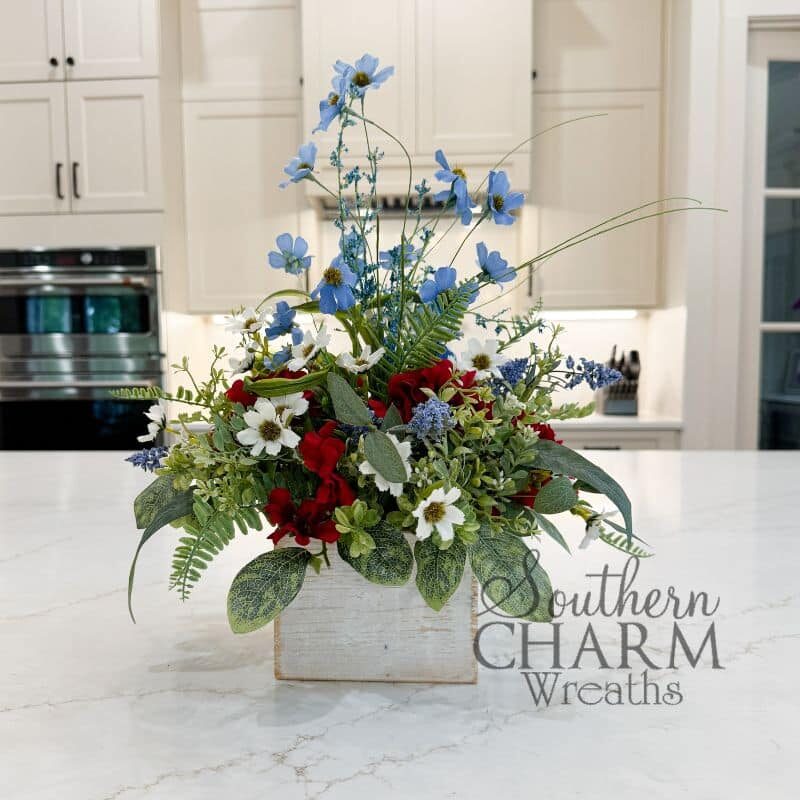 Patriotic table arrangement in a whitewashed wood box on a marble kitchen island. Features tall blue cosmos, red hydrangeas, white daisies, blue muscari, and mixed variegated greenery. Southern Charm Wreaths.
