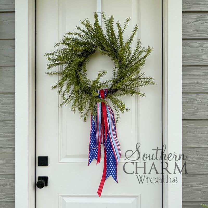 A full green fern wreath hanging on a white paneled front door with gray siding. A patriotic ribbon wreath sash made from three layered wired ribbons — navy blue with white stars, red and white stripe, and light blue and white stripe — is attached at the bottom center of the wreath and drapes in flowing tails below the base.