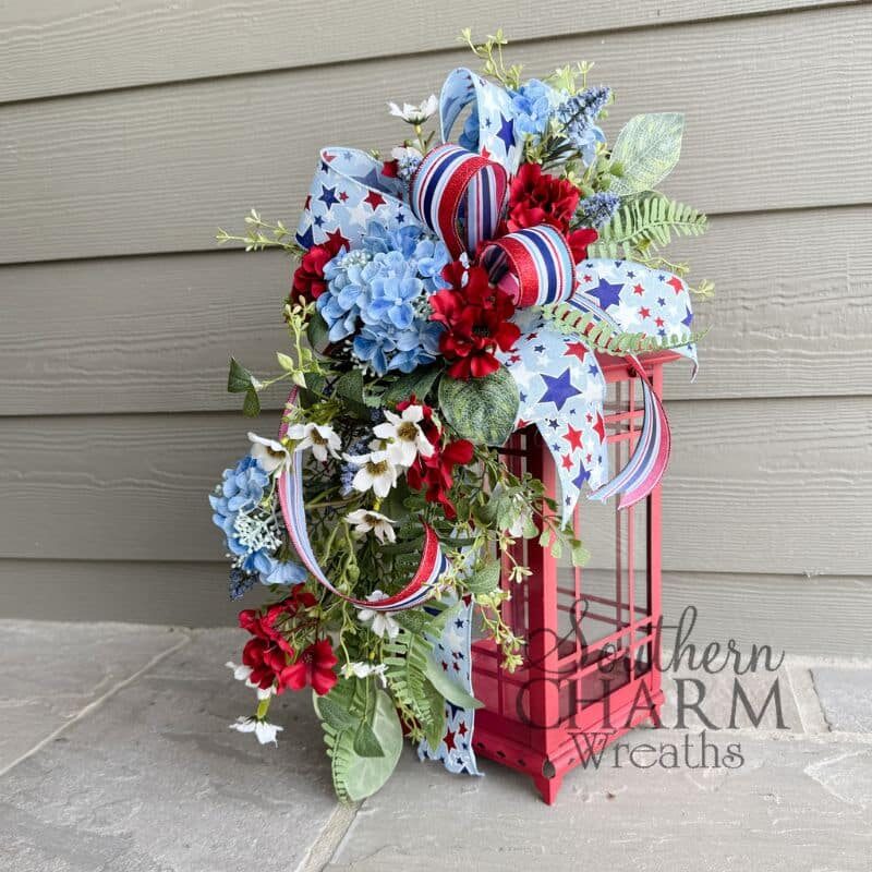Patriotic teardrop door swag with blue silk hydrangeas, red geraniums, white daisy filler, dusty lavender blooms, mixed greenery, and a layered star-print and red-white-blue striped wired ribbon bow.