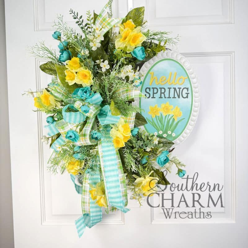 A vibrant spring daffodil grapevine wreath featuring yellow daffodils, teal roses, and a "Hello Spring" sign, hanging on a white door.