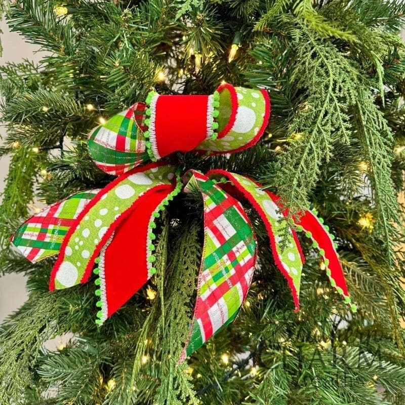 A festive Christmas tree bow made with bright red, green, and plaid wired ribbon, accented with pom-pom trim, perfectly nestled among lush greenery on a decorated tree.