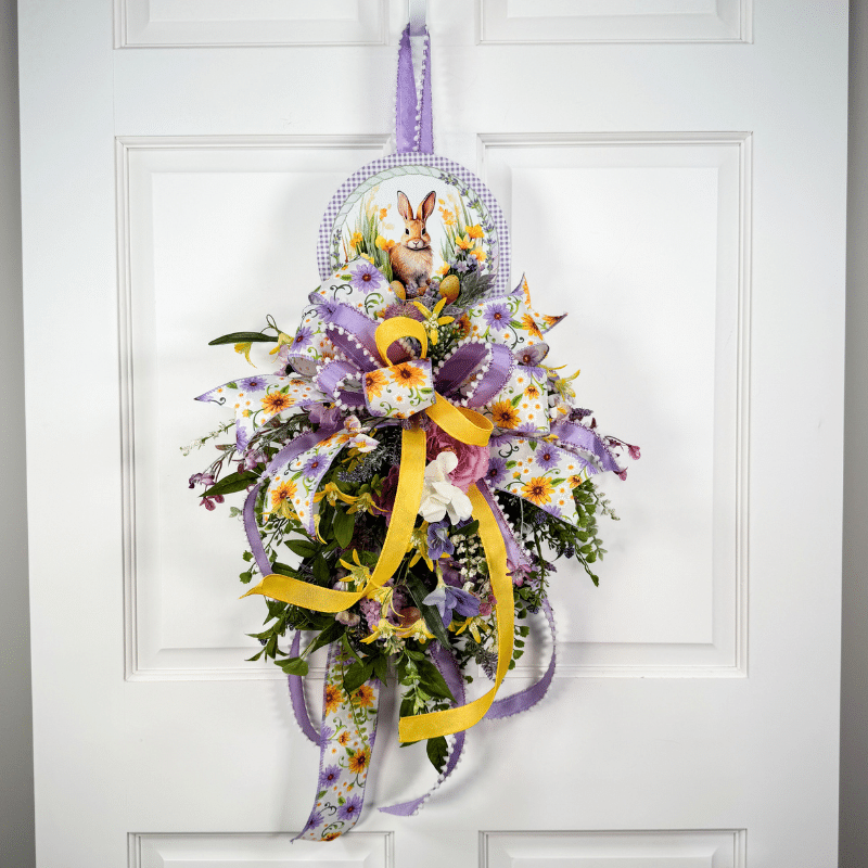 DIY Easter swag door hanger with bunny sign, purple and yellow ribbon bow, and layered spring florals arranged in a cascading vertical design on a white door.