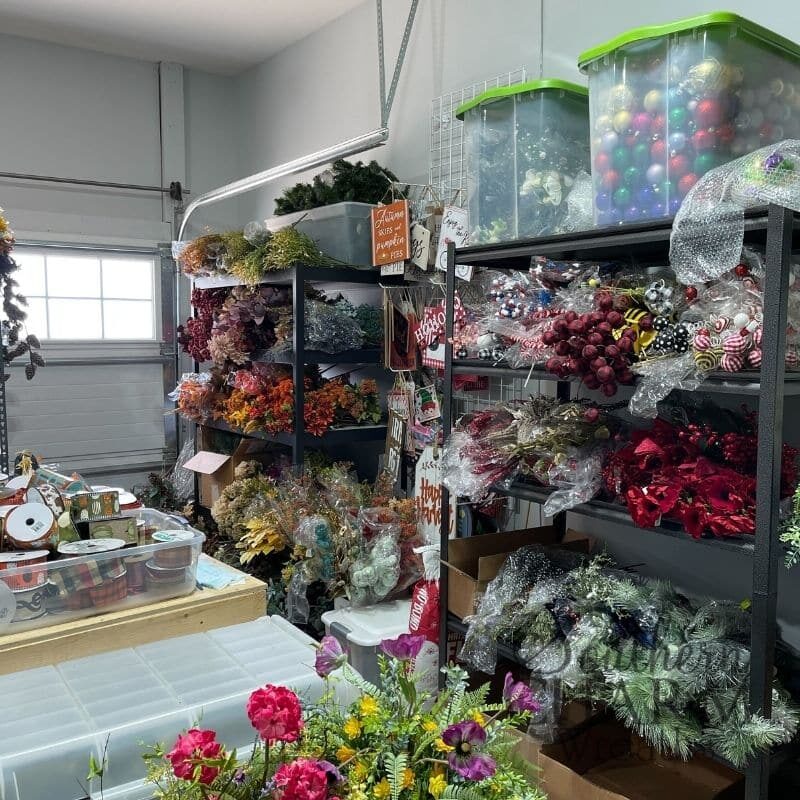 A garage transformed into a wreath shop with organized shelves of florals, ribbons, and craft supplies, perfect for DIY wreath making.